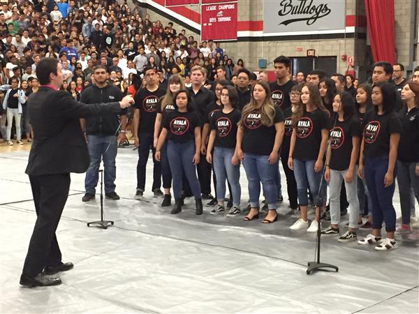 Members of the Ayala High Choir sing The National Anthem in a gym. They are directed by an adult teacher.